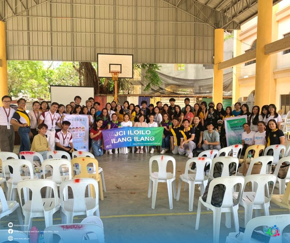 group of people in a covered basketball court posing for a picture; five people are holding a banner in the front and center, three people are holding a banner at the front left, and two people are holding another banner at the right; plastic chairs in rows with an aisle in the middle are facing the group of people